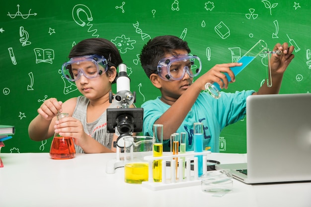 Photo kids and science concept - cute indian little kid student or scientist studying science or experimenting with microscope and chemicals with diagrams doodles drawn over green chalkboard