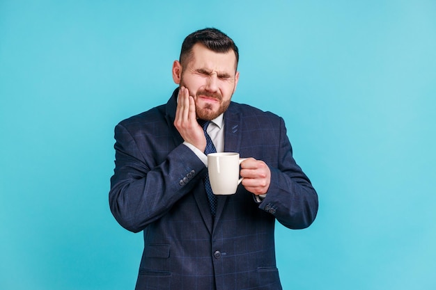 Kiespijn. Portret van een overstuur bebaarde man in een elegant pak die lijdt aan vreselijke tandpijn na het drinken van warme of koude dranken, tandletsel. Indoor studio opname geïsoleerd op blauwe achtergrond.