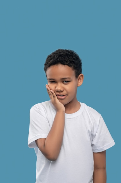 Kiespijn. Trieste Afro-Amerikaanse jongen in witte t-shirt die de wang aanraakt met palm op blauwe achtergrond in studiofoto