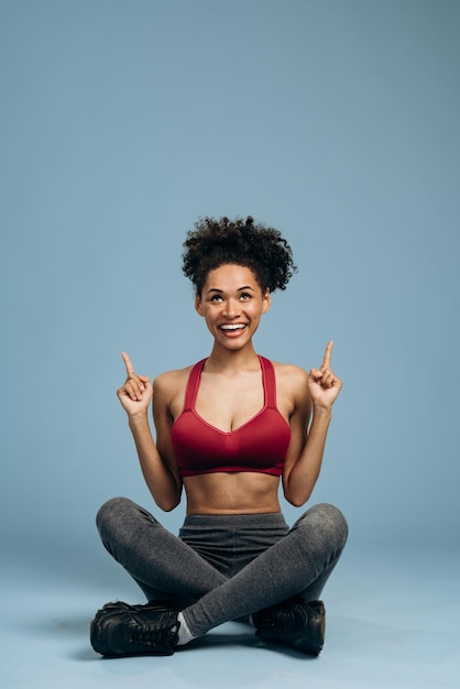 Kijk bij adverteren. Portret van een aangenaam verraste sportvrouw die naar boven wijst en met een blij gezicht kijkt, zittend in de lotushouding. Indoor studio shoot geïsoleerd