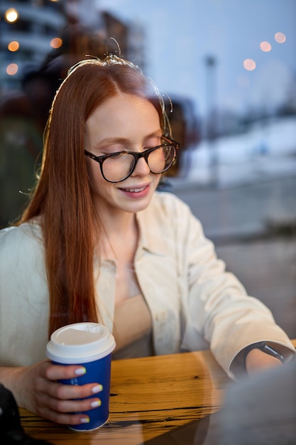 Kijk door het raam op roodharige vrouw met bril die in café aan freelance werkt. succesvol glimlachen in trendy outfit met laptop, tijd doorbrengen in coffeeshop