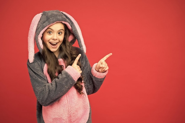 Kijk eens Gelukkig meisje wijzende wijsvingers naar iets Klein kind in konijnenpyjama wijzend op rode achtergrond Wijzend gebaar Wijzend en promotend Te koop kopieerruimte