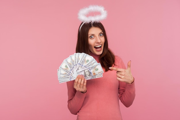 Kijk hiernaar. Opgewonden gelukkige vrouw met bruin haar en halo boven het hoofd wijzende vinger naar dollarbiljetten in haar hand, verrast met grote loterijwinst. Indoor studio-opname geïsoleerd op roze background