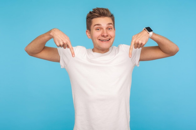 Foto kijk hieronder, plaats voor reclame. portret van een opgewonden, gelukkige man in een casual wit t-shirt dat naar beneden wijst en naar de camera kijkt met een vriendelijke brede glimlach. indoor studio-opname geïsoleerd op blauwe achtergrond