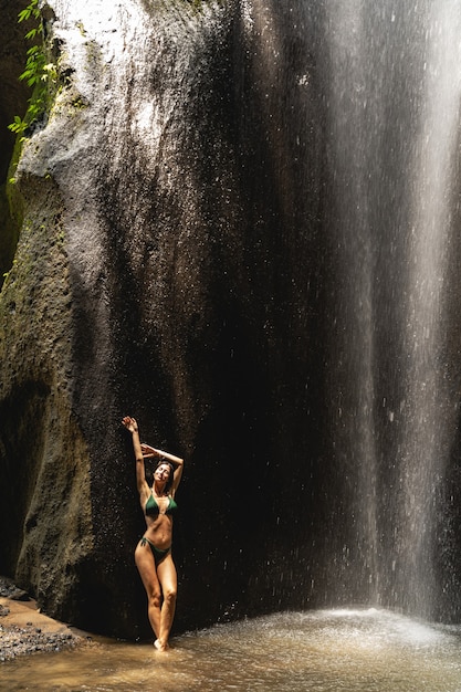 Kijk me aan. Vrolijke jonge vrouw die haar armen opheft terwijl ze voor de camera bij de waterval poseert