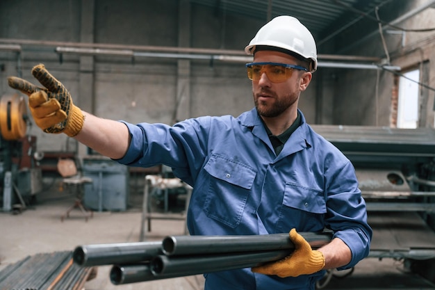 Foto kijk naar de zijkant en houd metalen leidingen fabrieksarbeider in blauw uniform is binnen