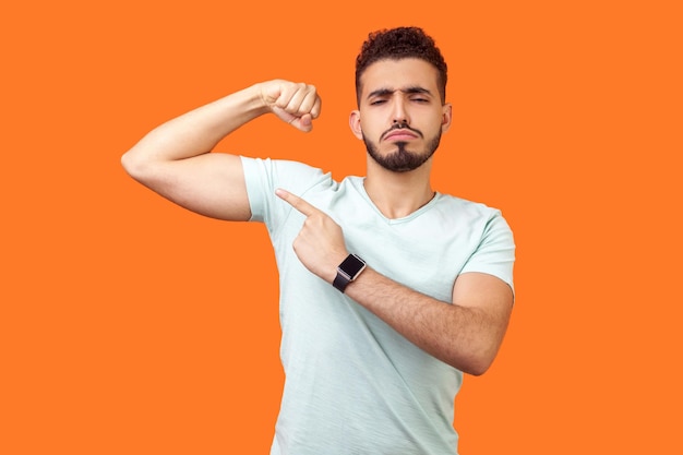 Kijk naar mijn kracht Portret van trotse knappe brunette man met baard in casual witte tshirt wijzend op biceps zelfverzekerd en krachtig indoor studio shot geïsoleerd op oranje achtergrond