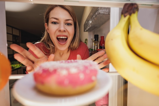 Foto kijk vanuit de koelkast naar buiten terwijl een vrouw de deur opent en opgewonden reikt naar een ongezonde donut. selectieve aandacht.