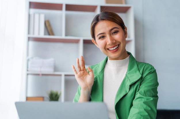 Kijken naar de camera Succesvolle jonge, mooie Aziatische vrouw die hallo zwaait tijdens een videogesprek bij het gebruik van een laptopcomputer aan een bureau op een kantoorwerkplek