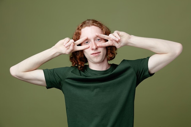 Kijkend naar de camera die vredesgebaar toont, een jonge knappe kerel met een groen t-shirt dat op een groene achtergrond wordt geïsoleerd?