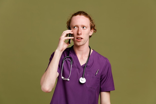 Kijkend naar de zijkant spreekt aan de telefoon jonge mannelijke arts in uniform met stethoscoop geïsoleerd op groene achtergrond