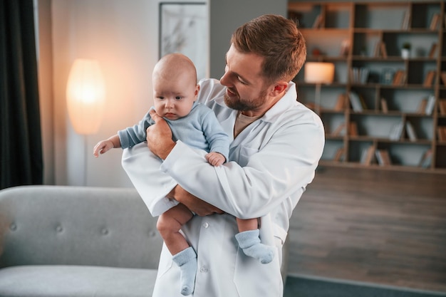 Foto kijkend naar het kind dokter staat en houdt pasgeboren baby in handen binnenshuis in het appartement