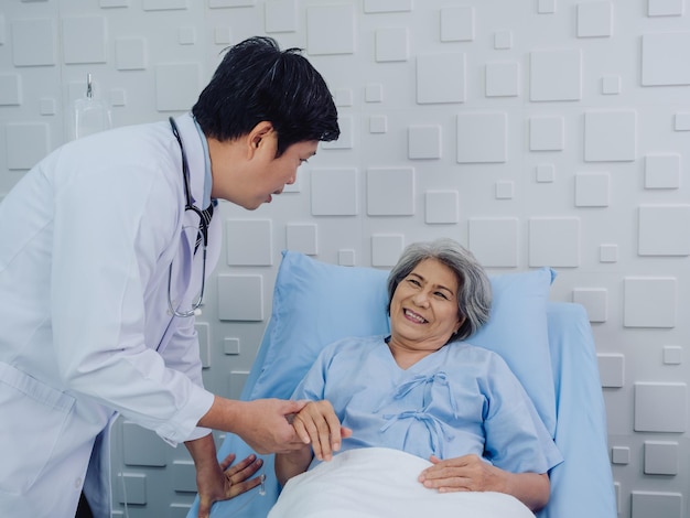 The kind Asian male doctor in white suit visits talks and gives support holding hand of happy elderly senior woman patient in light blue dress lying on bed in saline solution in hospital room