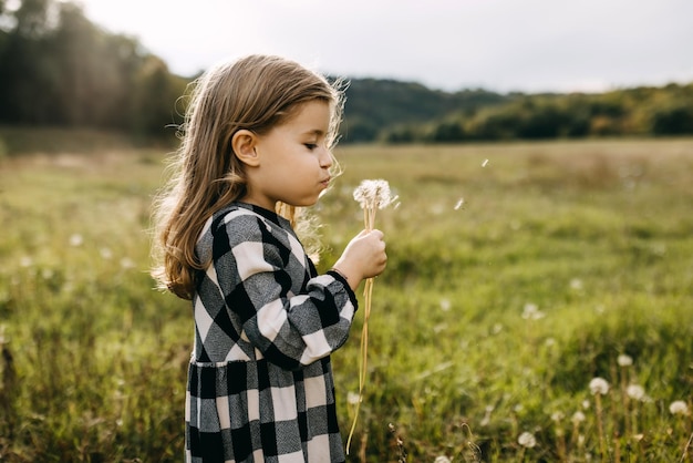 Kind blaast paardenbloemen die een jurk dragen op een zomerdag in een veld