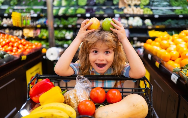 Kind dat een appel in een winkel kiest Grappige kleine kindglimlach en appel vasthoudt Kind winkelen in de supermarkt Jongetje met kar die verse groenten kiest in de plaatselijke winkel