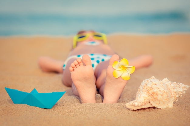 Kind dat op het zand op het strand ligt met een bloem aan zijn voet en een blauwe origamiboot en een schelp