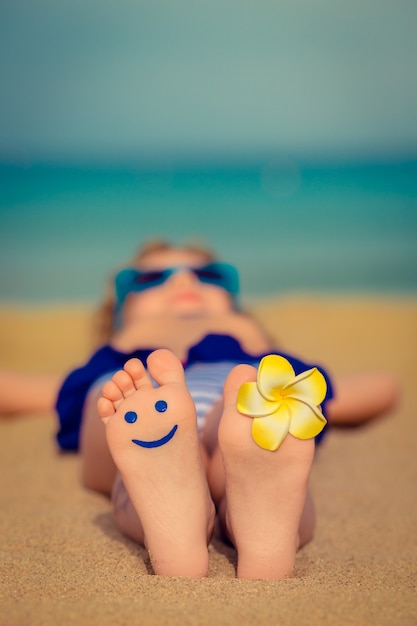 Kind dat op zandstrand ligt met een gele bloem op zijn voet en een smiley aan de andere kant