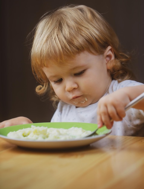 Kind dat voedingsconcept eet, schattig babygezicht met een lepel en een bord in de keuken thuis