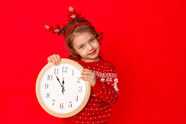 Kind een klein meisje in winterkleren houdt een horloge vast op een rode achtergrond Nieuwjaarsconceptruimte voor tekst