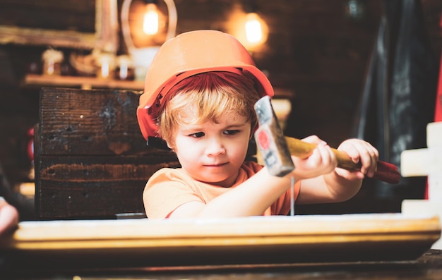 Kind hamert spijkers met een hamer in een houten plank Jongenskind bezig in helm die hamer leert gebruiken Kind die spijker thuis in hout hamert