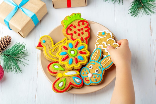 Kind houdt een zelfgemaakte geschilderde peperkoek (cookie) op witte houten achtergrond onder fir takken en geschenken. Kerstmis en Nieuwjaar zoet geschenk concept. Grappig zoet voedselclose-up.