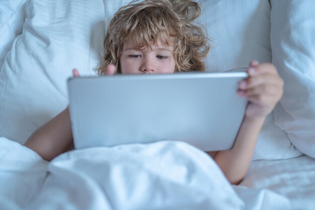 Kind in bed onder een deken speelt in de ochtend op een tablet het gezicht van het kind bij een tablet kind met behulp van ta