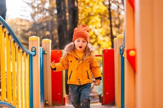 Kind in de herfst speelt gelukkig op de speelplaats in een gele parkjas en spijkerbroek