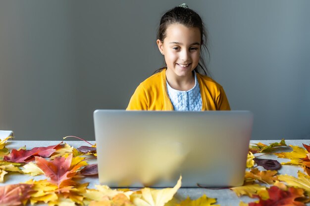 Kind in herfstbladeren met laptop en boeken.