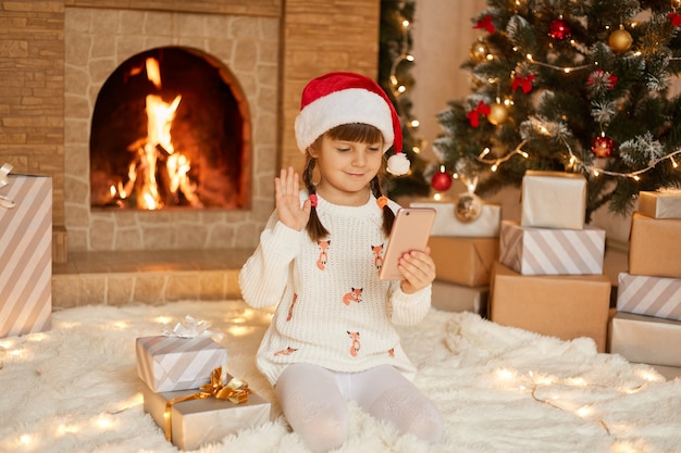 Kind in kerstmuts met web chatgesprek met iemand, scherm kijken en zwaaiende hand. Leuk peutermeisje met twee vlechten die de hoed van de Kerstman en witte trui dragen.