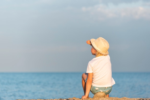 Kind in strooien hoed zittend op het strand en kijkt naar zee