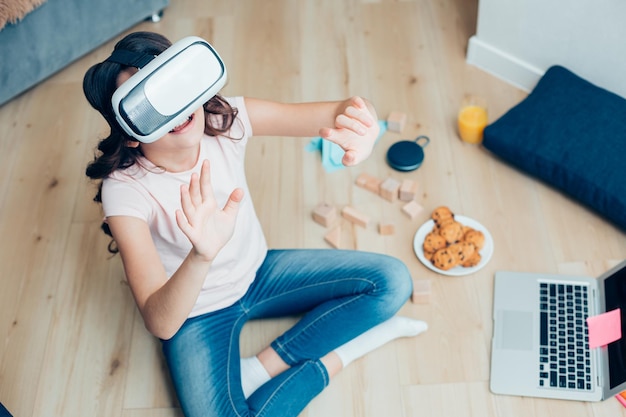 Kind in vrijetijdskleding zittend op de vloer met een laptop en een bord koekjes Virtual reality-apparaat op haar ogen