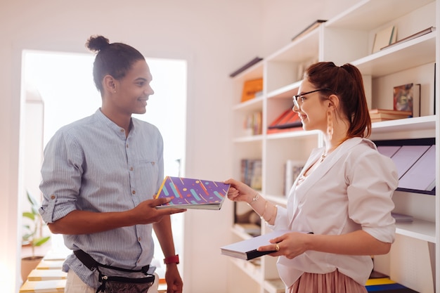 Kind international student keeping smile on his face while giving interesting book to pretty girl