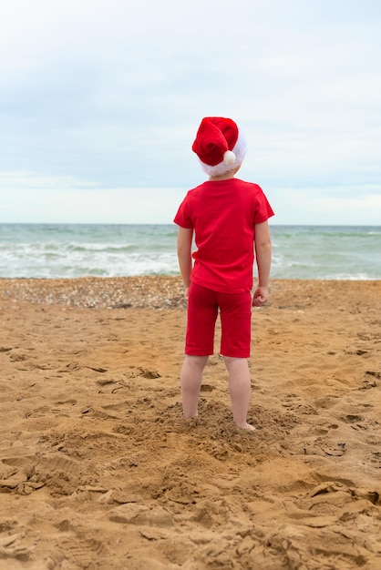 Kind jongen in rode doek en kerstman hoed genieten van Kerstmis op het strand op Xmas reizen vakantie Kerstmis of Nieuwjaar viering concept