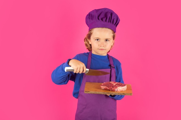 Kind kok houdt snijplank met vlees biefstuk en mes Koken culinair en kinderen Kleine jongen in koksmuts en schort op studio geïsoleerde achtergrond