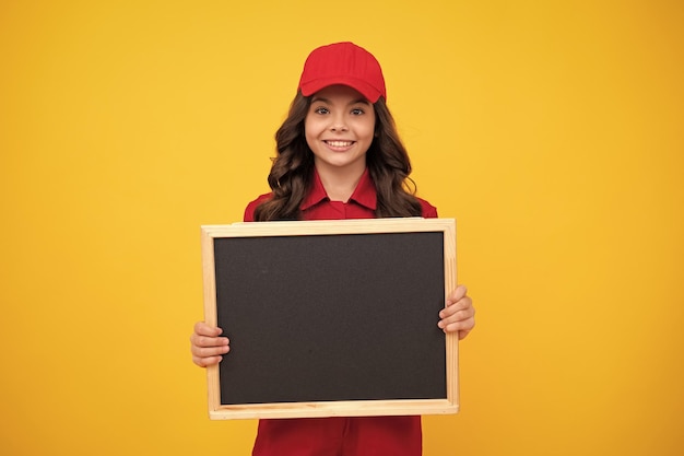 Kind meisje bouwer in rood gebouw uniform en cap tienermeisje werknemer houden schoolbord geïsoleerd op gele achtergrond Kids renovatie concept kopie ruimte op leeg bord mock up voor menu