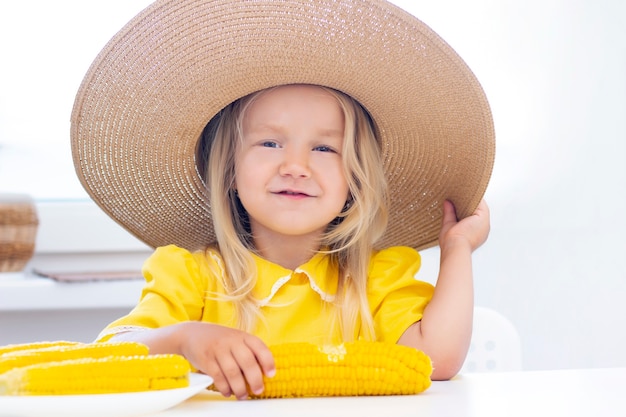 Kind meisje in een strooien hoed in gele kleren eet maïs