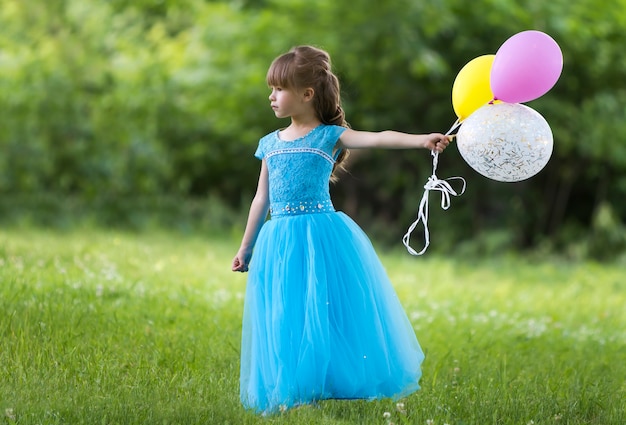 Kind meisje in mooie lange blauwe avondjurk houdt kleurrijke ballonnen.