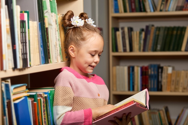 kind meisje laat zich meeslepen door het lezen van een boek in de bibliotheek, staande tussen veelkleurige boekenplanken