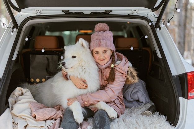 Kind meisje zit in de kofferbak van de auto met haar huisdier, een witte hond samojeed