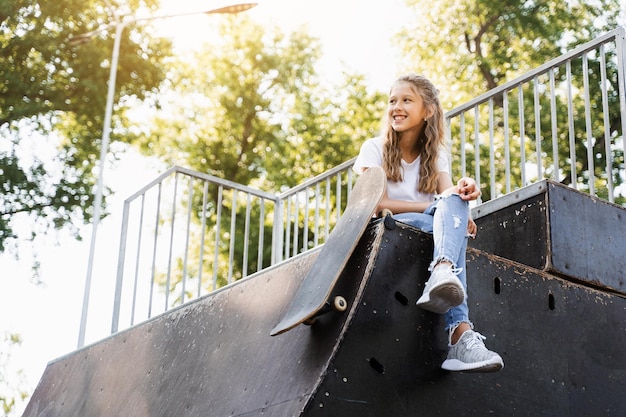 Kind meisje zit met skate board op sporthelling Sportuitrusting voor kinderen Actieve tiener met skate board op skatepark speeltuin