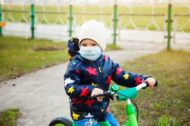 Kind op een ritje in een stadspark in een medisch masker rijdt op een fiets. Lopen op straat tijdens de quarantaineperiode van de coronavirus pandemie in de wereld. Voorzorgsmaatregelen en lesgeven aan kinderen.
