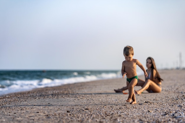 친절하고 마른 귀여운 누나는 여름 방학에 따뜻하고 화창한 날 해안 근처의 얕은 바다에 앉아 있는 동안 동생 유아와 놀고 있습니다.