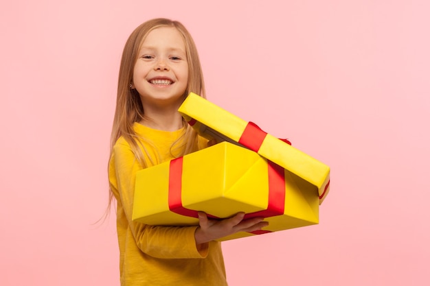 Kind tevreden met goed cadeau. Portret van een charmant, grappig meisje dat een geschenkdoos opent en naar de camera glimlacht, verjaardag viert, kerstvakantie. studio-opname geïsoleerd op roze achtergrond