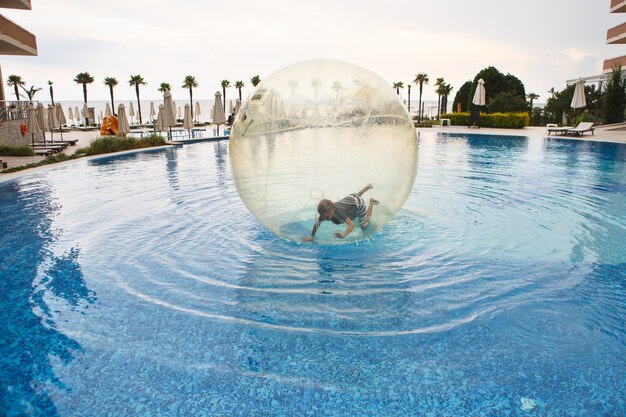 Kind veel plezier in grote plastic ballon op het water van het zwembad op het zomerverblijf. kleine jongen binnen grote opblaasbare transparante bal rennen en plezier maken.