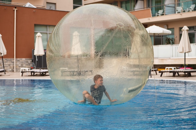 Kind veel plezier in grote plastic ballon op het water van het zwembad op het zomerverblijf. kleine jongen binnen grote opblaasbare transparante bal rennen en plezier maken.
