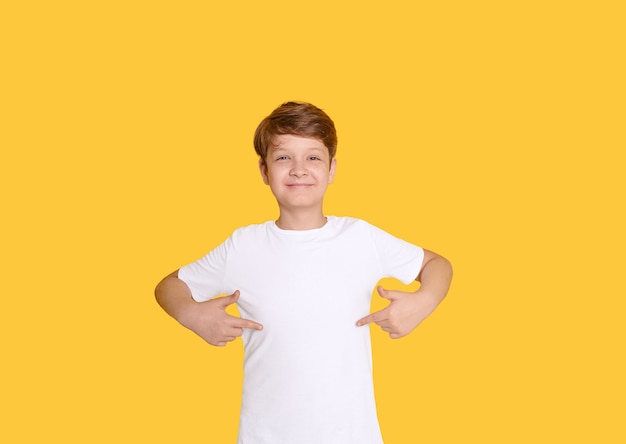 Kind wijst met zijn vingers op een leeg t-shirt, op gele achtergrond met een plek voor uw reclame.
