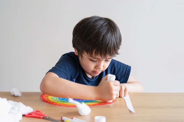 Kind zet lijmstift op papier voor het plakken van watten als decoratieve elementen voor wolken op regenboog