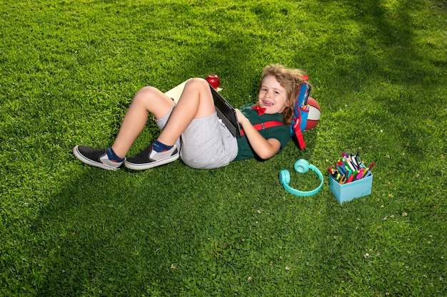 Kind zittend op gras met tablet en schoolbenodigdheden in het park Outdoor school