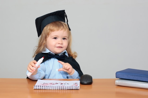 Kindbaby in academische mantel en GLB die bij de holdingsmarkering van het school houten bureau zitten