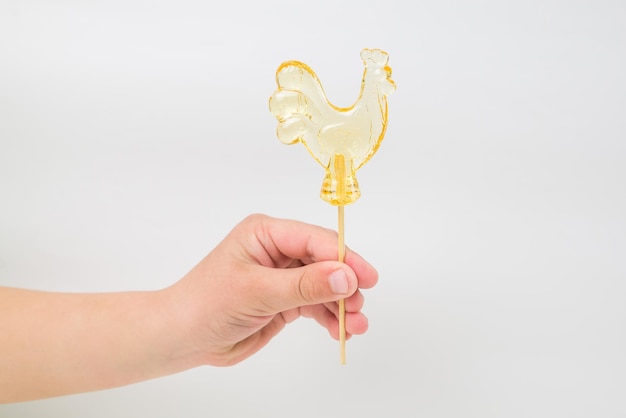 Kinder snoep karamel in de kindertijd Geïsoleerde oranje transparante lolly Cockerel op een houten stok in de hand op een witte achtergrond Vreugde herinneringen zoetheid geschiedenis